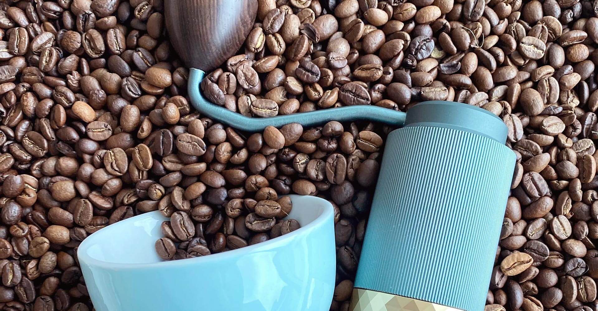 Blaue Handmühle und blaue Tasse liegen auf einem Bett aus Kaffeebohnen